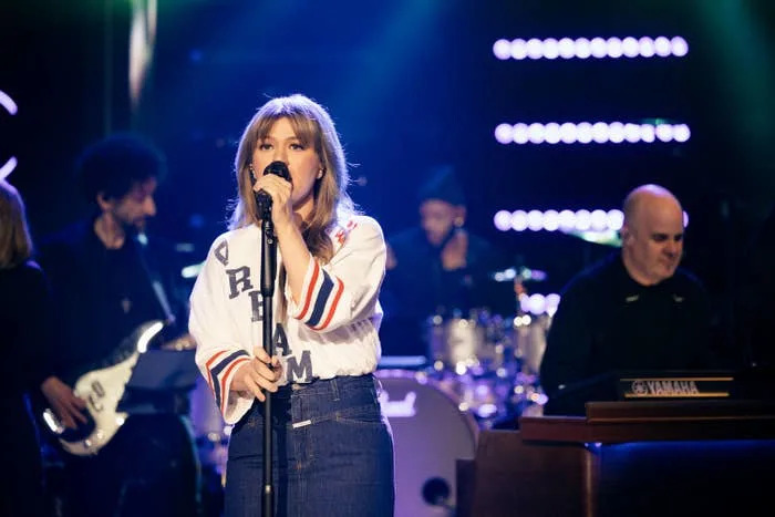 Kelly Clarkson performs on stage with band members, wearing a casual sports-style shirt and jeans, holding a microphone with stage lights in the background