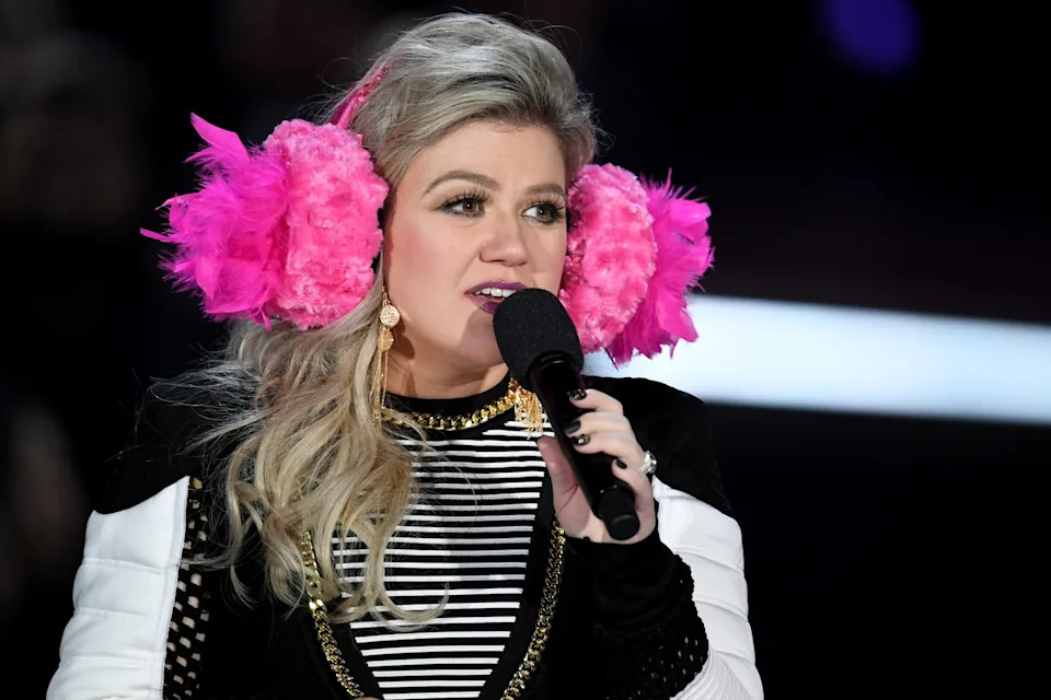 Kelly Clarkson with oversized ruffly earmuffs speaks into a microphone, wearing a striped top with gold accents