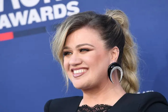 Kelly Clarkson smiling on a red carpet, wearing an outfit with lace detail and bold crescent-shaped earrings