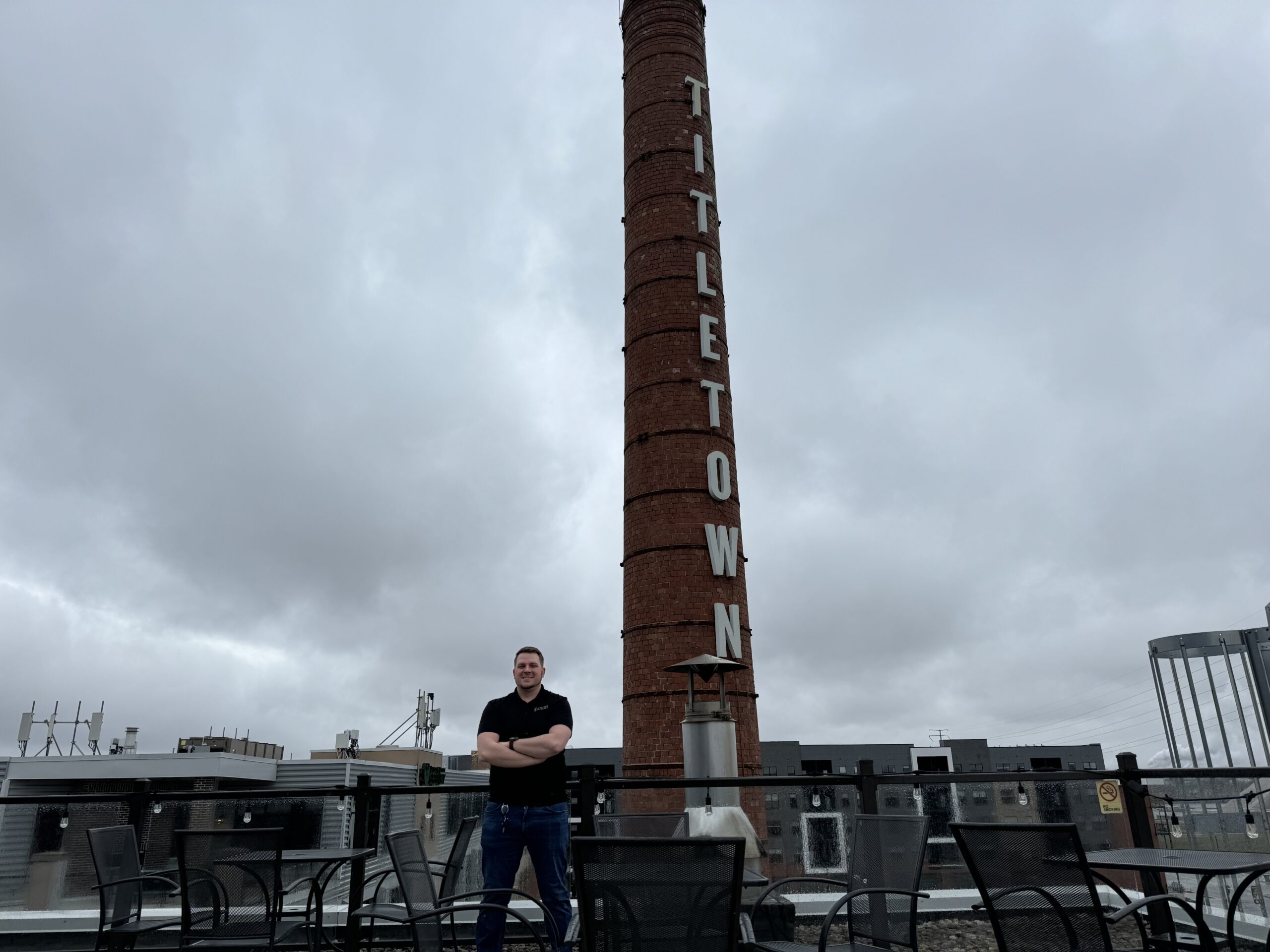 A man stands with arms crossed in front of a tall brick smokestack labeled TITLETOWN on a cloudy day, surrounded by empty outdoor chairs and tables.