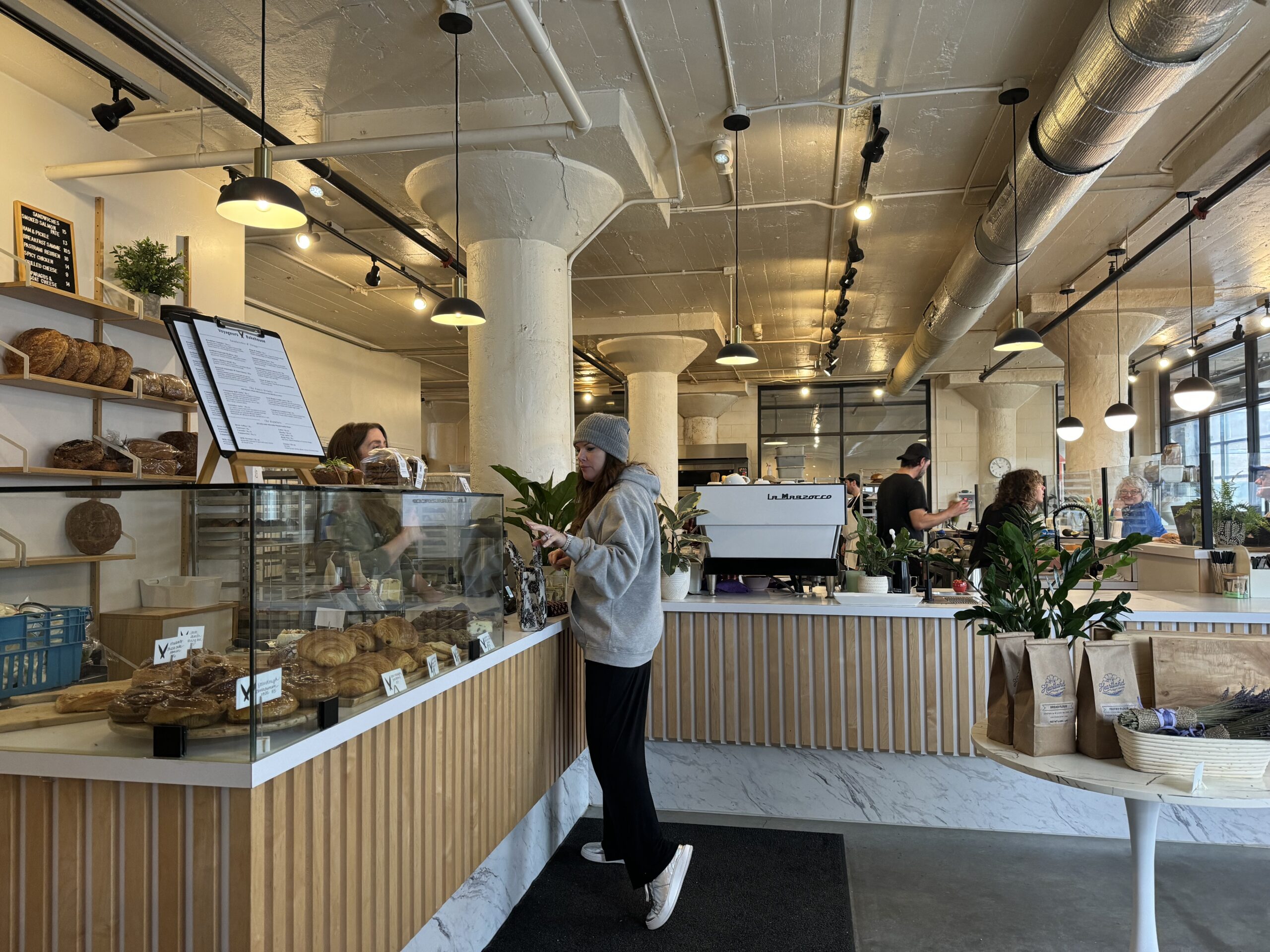 Several people are inside a modern bakery cafe; a woman in a gray sweatshirt stands at the counter near a display of pastries and a menu, while staff work behind the counter.