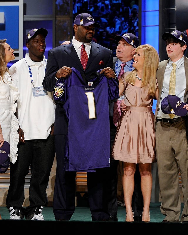 michael oher stands in a group of people while holding up a baltimore ravens jersey, he wears a suit and a baltimore ravens hat, his family are dressed in business formal clothes