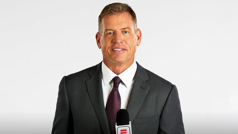 Troy Aikman standing with a ESPN microphone on a white backdrop