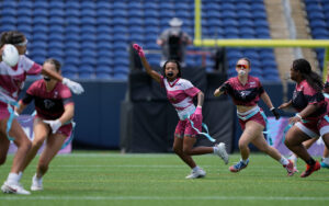 The image depicts a group of female athletes playing flag football on a green field within a stadium. The players, wearing maroon and white outfits with light blue belts holding flags, are actively engaged in the game.