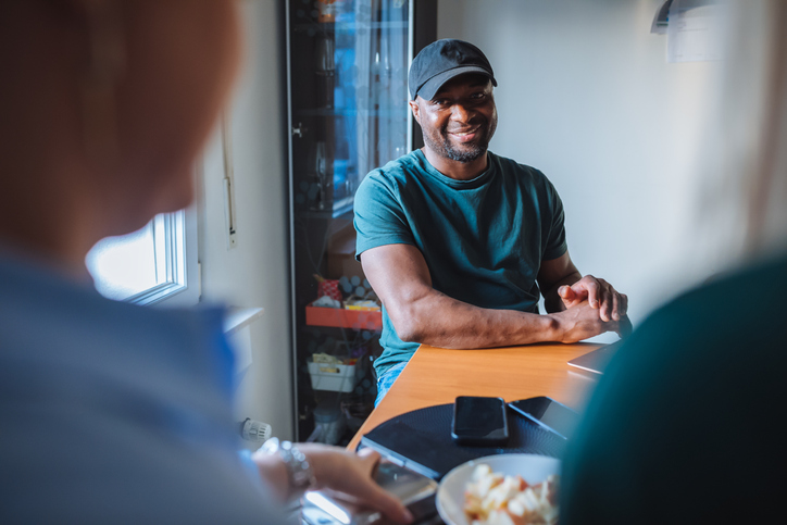 Happy black man resting at kitchen table and looking at camera, smiling brightly, posing between two unrecognizable white women sitting on foreground in front of male.
