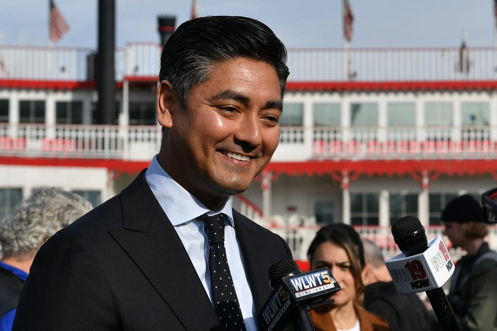 cincinnati major aftab pureval smiles as unseen people hold microphones toward him, he wears a suit and tie and stands outside