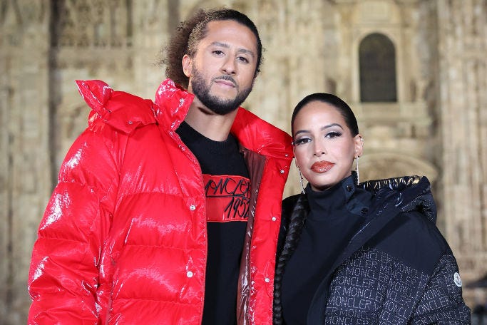 colin kaepernick and nessa diab pose for a photo while standing outside a cathedral, he wears a black shirt with red detailing and a red puffy coat, she wears a black turtleneck with a black puffy coat