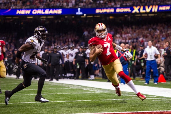 colin kaepernick runs on a football field just outside a red endzone while holding a football and wearing a san francisco 49ers uniform, a player for the baltimore ravens runs toward him from the left, ravens players and staff stand in the background and fans watch from stands