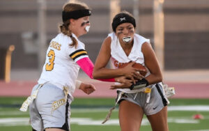 Two players in action during a flag football game, one holding a football and the other reaching towards her.
