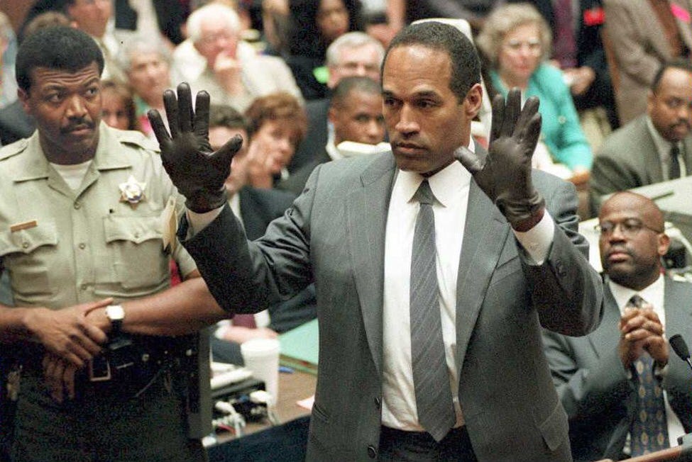 oj simpson raises both of his hands in the air in front of him and looks left, he wears black gloves and a gray suit and tie with a collared white shirt, many people look on from the background