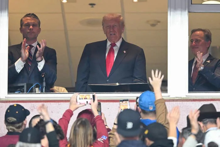 A man in a suit and red tie stands in a booth, attended by two others, as a crowd below watches and takes photos