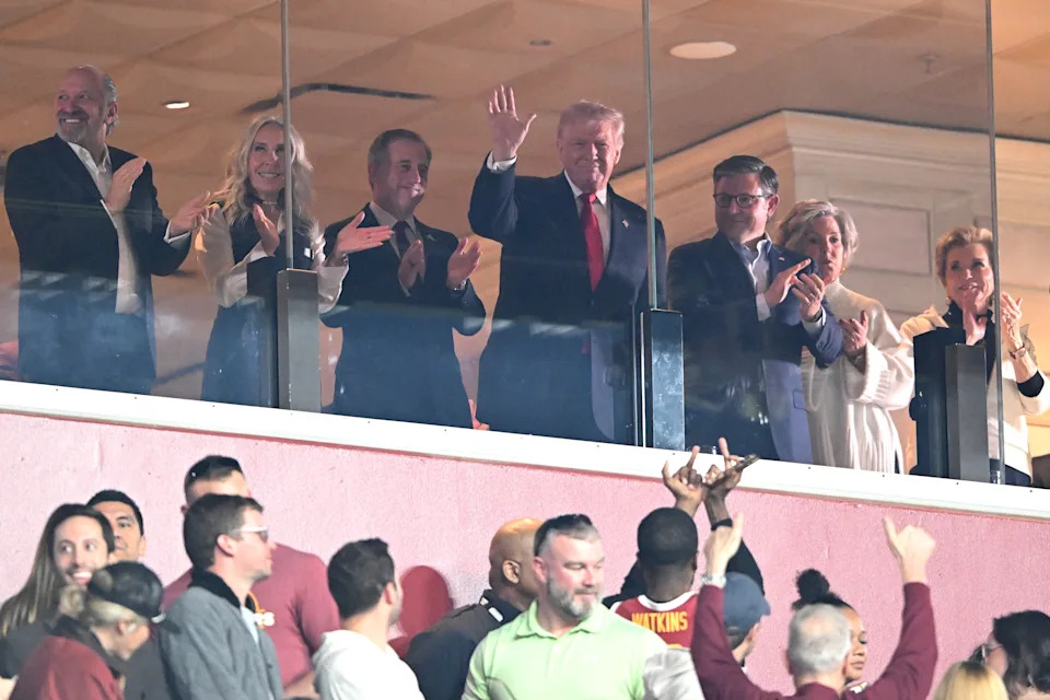 A group of people, including a man in a suit waving, stand in a sports box and applaud while spectators below look and cheer